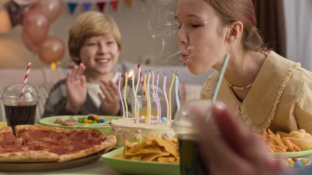 Caucasian preteen girl sitting at table, closing eyes and blowing out candles while making wish. Her friends looking ather and clapping hands