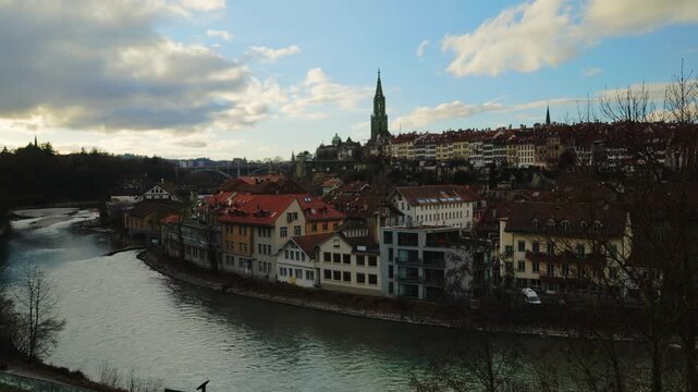 The towering gothic spire of the Bern Minster rises above the historic riverfront houses and the emerald waters of the Aare, captured under a dramatic canopy of gray clouds that emphasize the timeless