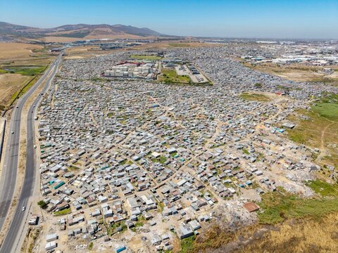 Aerial view of corrugated iron roofs blanketing the terrain, stark against the pale earth and distant mountains, a visual echo of human resilience, Cape Town, Western Cape, South Africa.