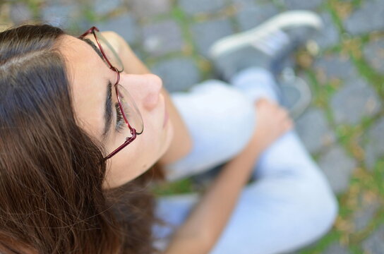 Overhead view of a teenage girl with glasses wearing white jeans sitting on the pavement