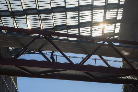 View of a solitary figure traversing a red steel pedestrian bridge against the glaring sun under a geometric glass roof, Punggol Coast, Singapore.