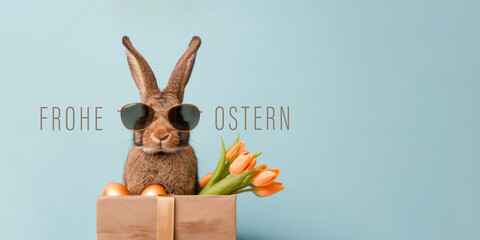 Frohe Ostern Konzept Feiertag Gru&szlig;karte mit deutschem Text - Cooler Osterhase, Kaninchen mit Sonnenbrille, sitzt in Geschenkbox mit Ostereiern, isoliert auf blauem Hintergrund