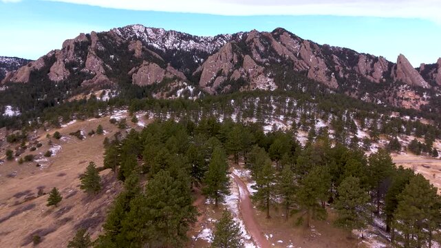 Aerial drone footage flying over a snowy hiking trail in the Colorado foothills toward the Flatirons sandstone rock formations in a scenic winter mountain landscape