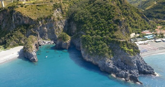 Aerial view of a secluded coastal cove featuring deep turquoise waters nestled between steep, rugged cliffs and a lush, green-covered mountain, with a small beach and a boat visible. Italian coast
