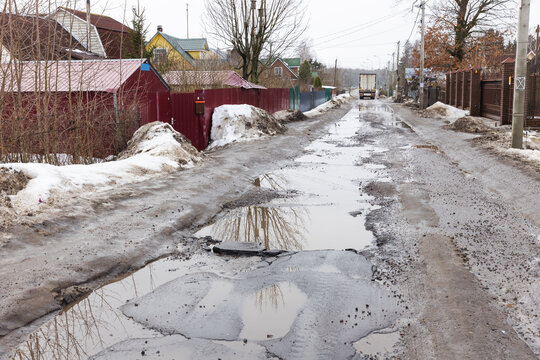 A muddy, puddled rural street lined with fences, bare trees and snow piles