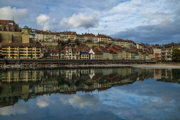 Fototapeta premium The historic facades and warm-toned rooftops of Bern's riverside district are perfectly mirrored in the tranquil, emerald waters of the Aare river under a vast sky of soft, rolling clouds.