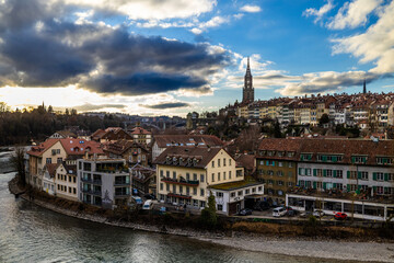 Obraz premium The historic houses of the Matte district line the banks of the turquoise Aare river, while the majestic spire of the Bern Minster rises above the city’s rooftops under a dramatic, cloud-filled aftern