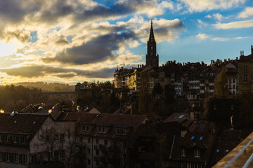 Obraz premium The dramatic silhouette of the Bern Minster dominates the skyline as golden evening light breaks through heavy clouds, illuminating the rooftops of the old city and the distant arched bridge.
