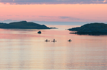 Fototapeta premium Midnight sun, evening, night. Amazing pink, orange sky, with reflection in the water of the sea, ocean. Some small rocky islands. 3 kayakers in the sea enjoying the light. Adventure, sports during mid