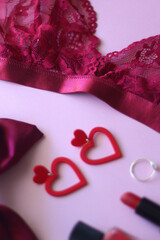 Red lace bralette, hairbow, heart shaped earrings, golden ring, red lipstick and nail polish on pink background. Coquettish accessories for Valentine's day. Selective focus.