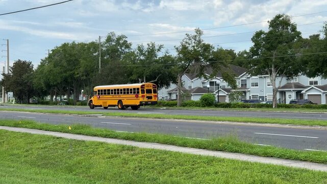 A bright yellow school bus drives past homes in a suburban neighborhood. Green trees and a yellow fire hydrant line the road on a cloudy day.