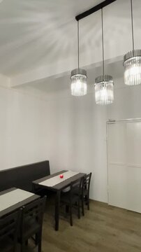 Crystal Chandelier Casting Patterned Ceiling Light, Long Bench Table And Chairs, Wooden Floor, White Walls, Minimalist Dining Interior With Soft Taken on mobile device.