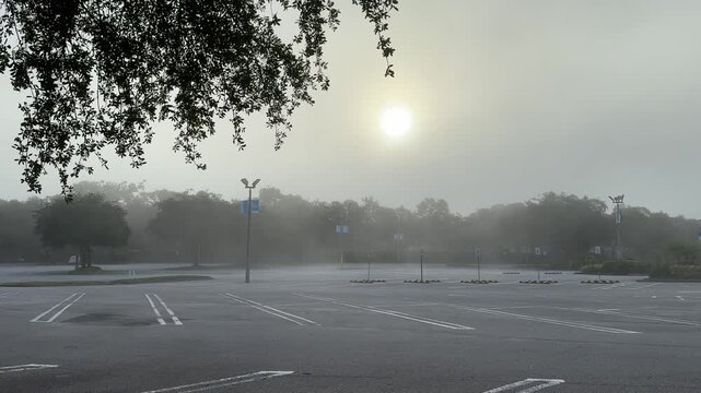 A large empty parking lot lies under a blanket of early morning fog. The rising sun glows softly through the mist above distant trees.