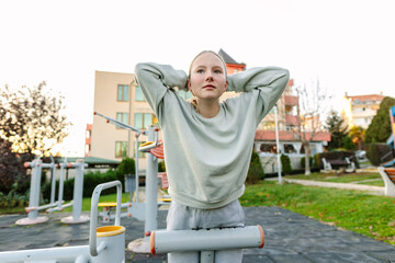 Teenage girl stretching and resting after workout on outdoor fitness equipment in park, healthy lifestyle, wellness, active childhood concept
