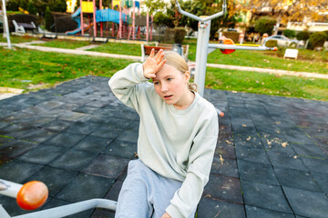 Teenage girl resting after outdoor workout on public fitness equipment, tired but calm expression, recovery after exercise, healthy lifestyle concept
