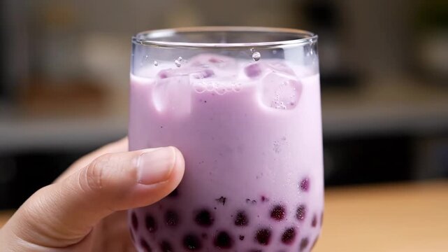 Iced Taro Bubble Tea Drink - This video showcases a person holding a glass of iced taro bubble tea. The drink contains tapioca pearls at the bottom with a creamy, lavender-colored liquid and ice