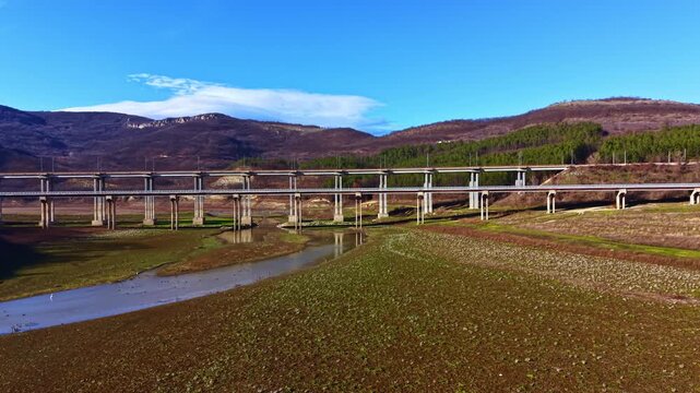 A bridge stands tall across a wide valley with dry ground and a small river below. The hills in the background have patches of trees and a clear blue sky above.