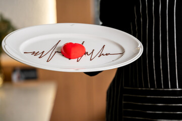 A chef wearing a striped apron presents a white plate featuring a heart-shaped red dessert and a drawn chocolate electrocardiogram (ECG) line