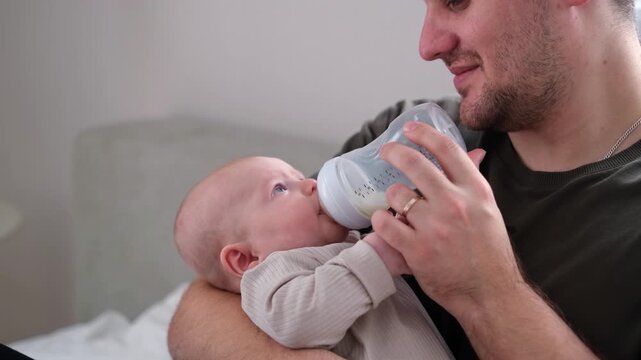 A father feeds his newborn son from a bottle of milk while sitting on the bed. The concept of caring for infants by fathers