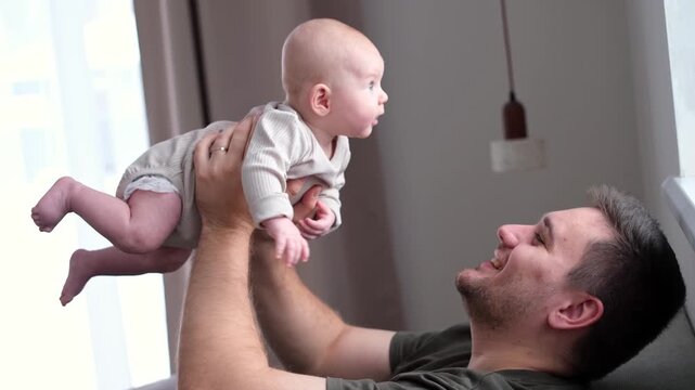 The middle plan of the portrait is in warm colors of a young father and newborn son. A father plays and kisses his little son on the bed. The concept of love, happy fatherhood.