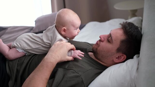 The middle plan of the portrait is in warm colors of a young father and newborn son. A father plays and kisses his little son on the bed. The concept of love, happy fatherhood.