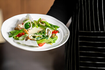 A waiter holds a white plate of fresh seafood salad with squid rings, mussels, tomatoes, and greens in a professional restaurant setting