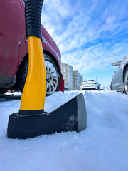 An axe with a yellow handle stands in the snow next to a red car