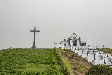 View of the historic Our Lady of Peace Chapel in Vila Franca do Campo, featuring its unique decorated staircase and a large stone cross under a misty sky