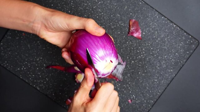 Hands peel a red onion in a kitchen setting during daytime. A person uses their hands to peel a red onion on a dark cutting board in a kitchen.