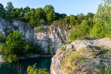Sunlit limestone ledge above zakrzowek quarry lake in krakow, with scrub oak and birch saplings growing between rocks, and dark turquoise water below creating a rugged nature and geology background.