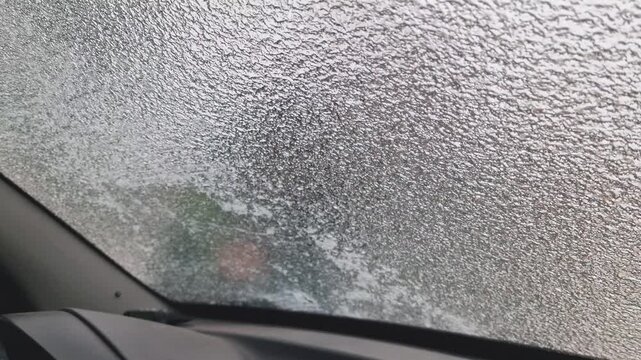 POV from inside a car: person uses a hand scraper to remove thick ice and sleet from the windshield on a cold winter day. Detailed texture of frozen glass being cleared for safe driving.