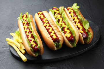 Tasty hot dogs with ketchup, mustard, lettuce and potato fries on black table, closeup