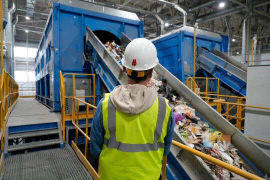 Male worker in a reflective vest and white helmet against the background of waste processing and sorting equipment on a conveyor belt, monitoring the quality of work of employees and personnel.