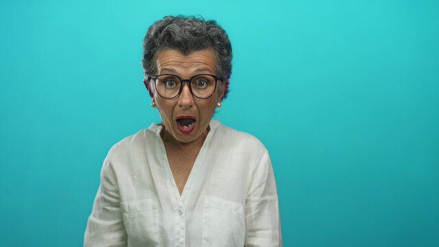 Senior woman with grey hair and glasses looks surprised against blue background, wearing white shirt, conveying shock and emotion in this vibrant studio setting.