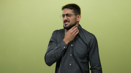 Obraz premium Young man wearing glasses in discomfort gestures against a green background conveying stress or throat pain in an isolated indoor setting for health or lifestyle concepts.