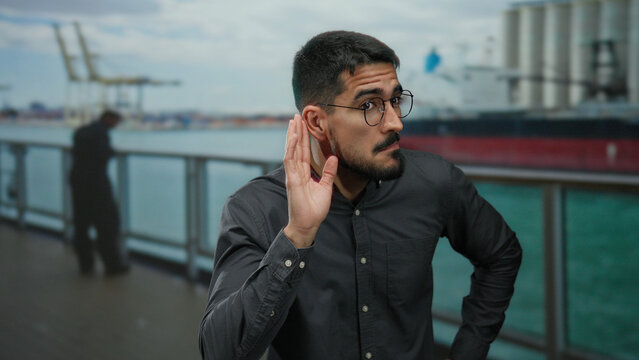 Hispanic man making a listening gesture by the seaside at a port with ships, highlighting an outdoor maritime environment.