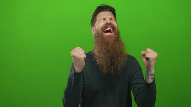 Man with long beard and tattooed forearm pumps fist to camera with smiling expression in green studio; excitement.