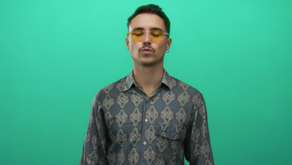 Fototapeta premium Young man with patterned shirt and yellow glasses standing against a vibrant green background looking confident and stylish in an isolated setting.