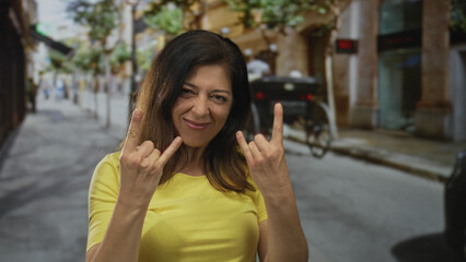 Fototapeta premium Middle aged woman makes hand rock horns gesture with open mouth expression on city street; rock fun metal energy.