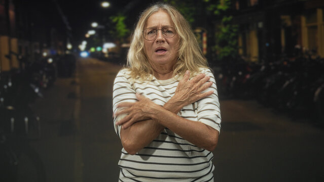 Senior woman with folded arms and pursed lips standing under streetlights on a quiet urban street at night with hunched shoulders and bowed head; vulnerability solitude.