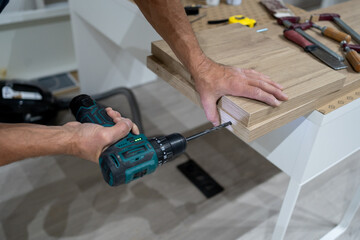 Carpenter drilling a hole in a plywood board using a cordless drill. Furniture assembly process in a home workshop, DIY and woodworking concept.