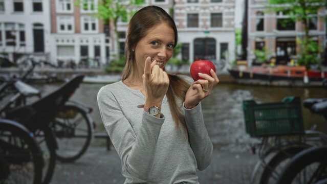 Woman holding red apple and beckoning with fingers on a street in amsterdam; playful healthy invitation.