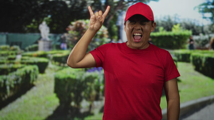Man in red cap and shirt making rock horns gesture in park with green hedges and a distant statue near walkway; playful joy youthful energy.