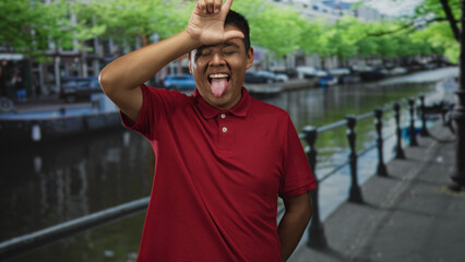 Fototapeta premium Man wearing red polo sticks out tongue and forms l with hand to forehead on street by canal in amsterdam; playful mischief.