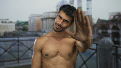 Obraz premium Young hispanic shirtless man raising palm as stop sign before roman ruins building; refusal defiance determination.