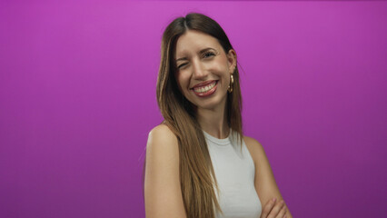 Fototapeta premium Woman with hoop earrings and long brown hair wearing a white tanktop smiling against a purple studio wall; confidence.