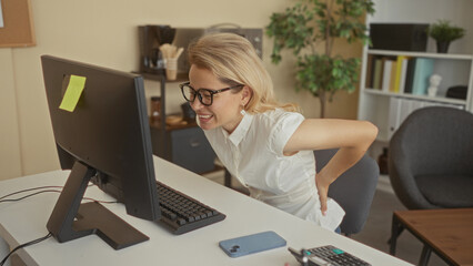 Obraz premium Woman wearing glasses typing on keyboard and holding lower back in pain in office building; stress backache.