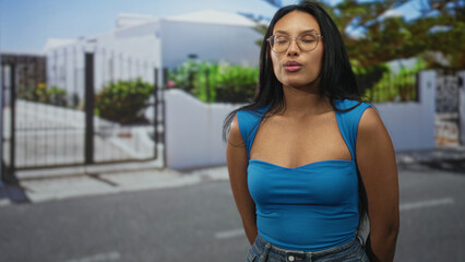 Obraz premium Woman with exposed chest and pursed lips wearing glasses standing on a street by a gated house; quiet contemplation.
