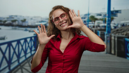 Fototapeta premium Smiling woman in a red shirt and glasses enjoying the seaside promenade with a joyful demeanor against a background of water and blue railings at the beach.