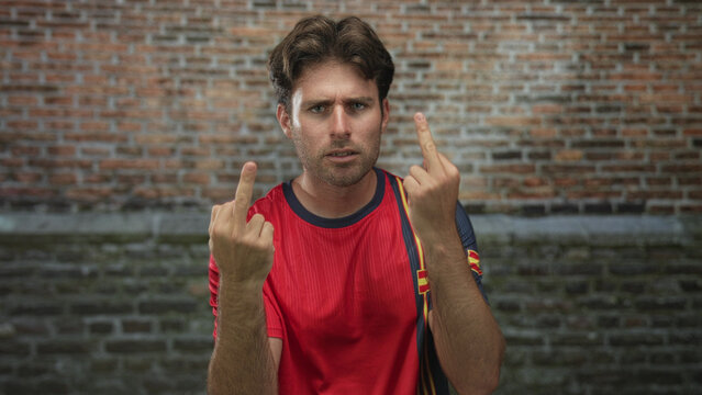 Man wearing red soccer jersey shows middle fingers in front of brick building, raising both hands in an aggressive gesture; angry defiance.
